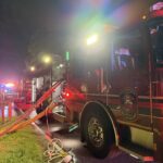 A fire truck with hoses attached is parked at night with emergency lights flashing. Other emergency vehicles and firefighters are visible further down the road. Grass lines the roadside.