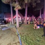 Firefighters in full gear stand outside a house at night, with smoke visible from the roof. Fire trucks and hoses are present, and emergency personnel are coordinating on a wet driveway near several tall trees.