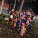 Three firefighters in full gear stand on grass at night near trees, with a residential house and emergency equipment visible in the background.